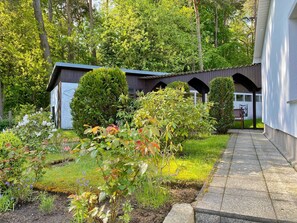 A garden with a pathway leading to a building with a dark facade and a white trim. There are various shrubs and a small tree in the foreground.