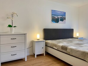 Um quarto com uma cama, mesas de cabeceira com luminárias, cômoda, planta em vaso e um quadro na parede.