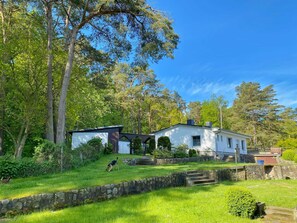 Uma casa com parede de pedra e pequena varanda, rodeada por vegetação exuberante e árvores.