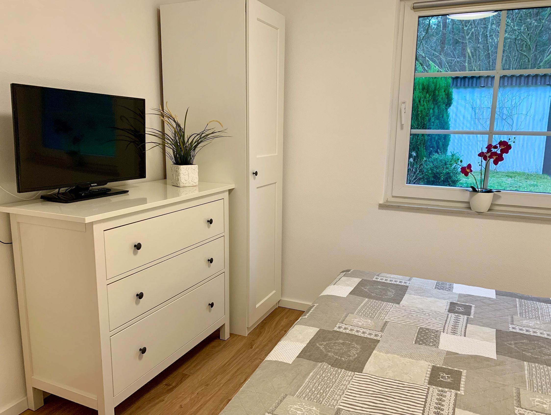 A bedroom with a bed, a TV on a dresser, a window with a view of greenery, and a potted plant on the windowsill.