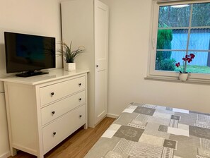 Een slaapkamer met een bed, een televisie op een dressoir, een raam met uitzicht op groen, en een potplant op de vensterbank.