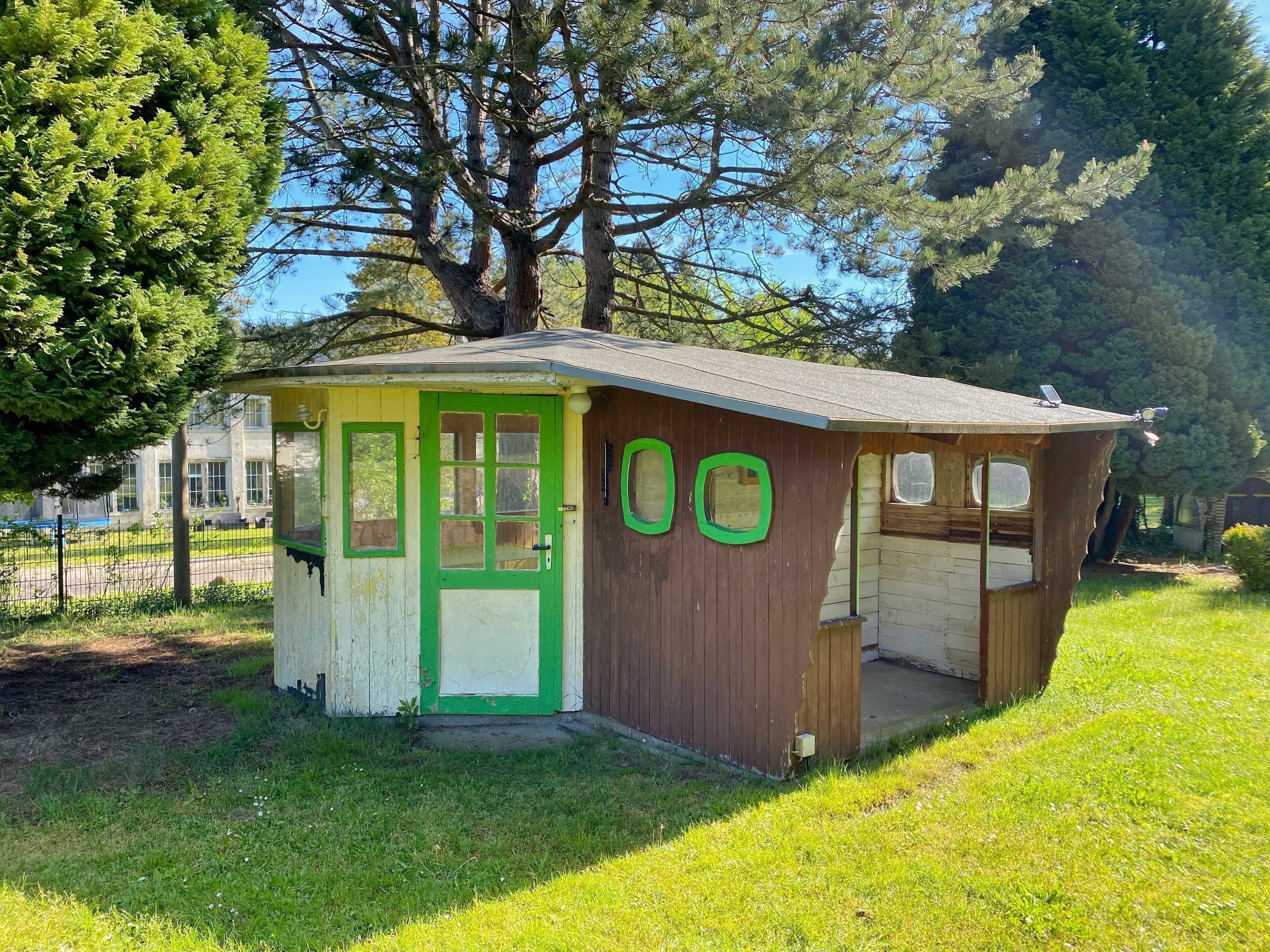 Una pequeña estructura de madera con puerta y ventanas verdes, rodeada de hierba y árboles.