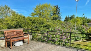 Une terrasse en bois avec un banc et un conteneur blanc, entourée de verdure et de fleurs violettes.