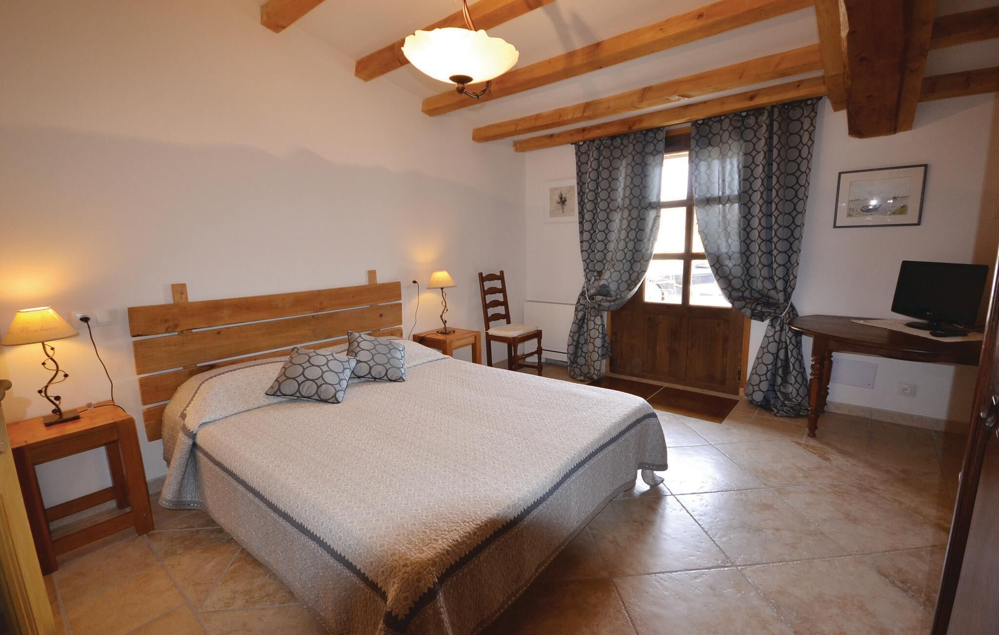 A bedroom with a wooden bed, two bedside lamps, a chair, a desk with a computer, and a window with curtains.