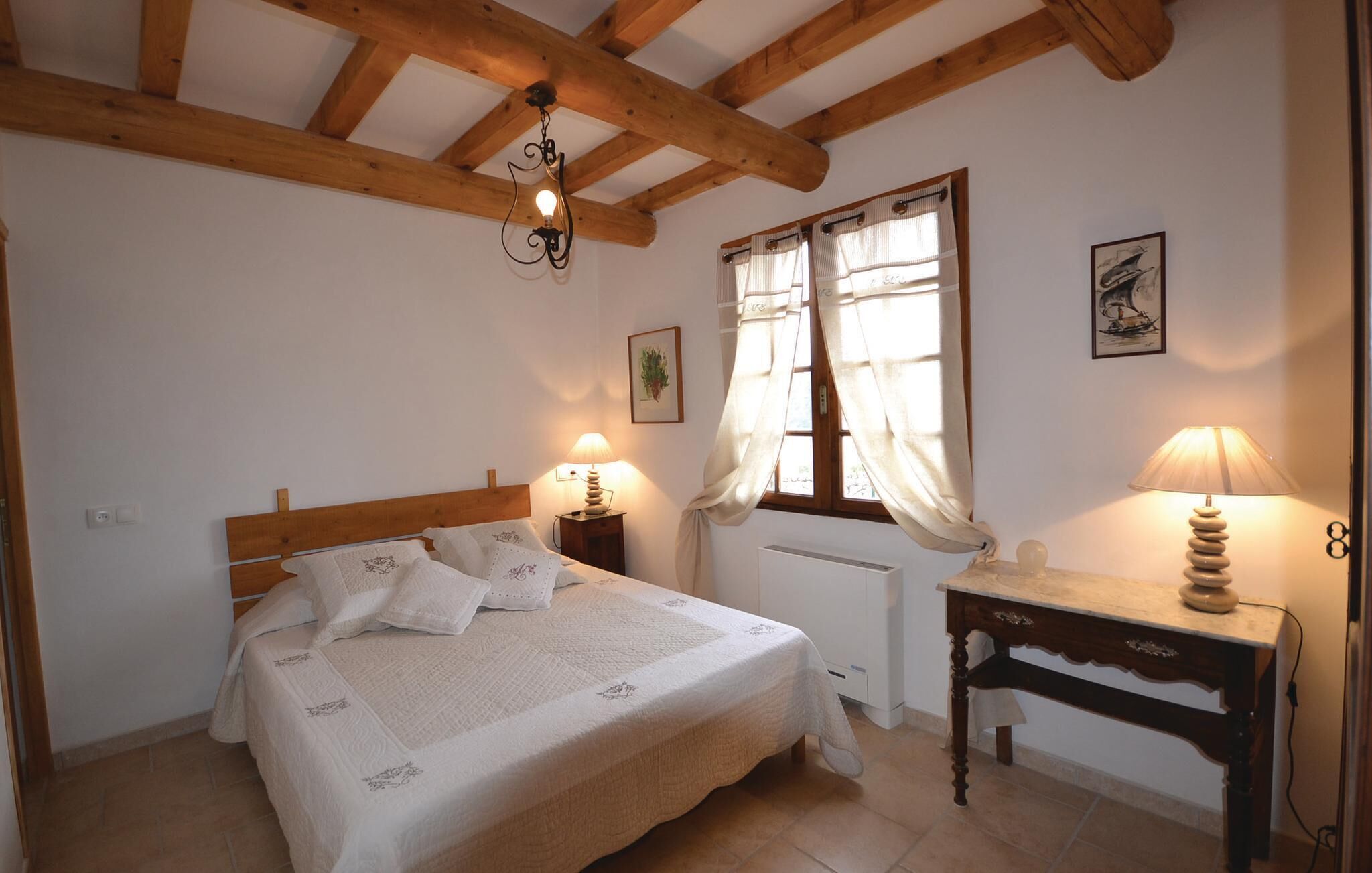 Ein Schlafzimmer mit Holz-Kopfteil, einem Bett mit weißer Bettwäsche, einem Nachttisch mit Lampe, einem Fenster mit Vorhängen und einem Heizkörper.
