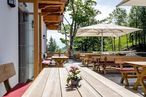 Een terras met houten tafels en stoelen, een plant in pot en een grote parasol.