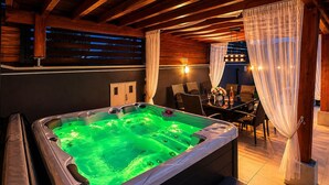 Een hottub met een groen licht, houten balkenplafond en een eethoek met stoelen en een tafel.