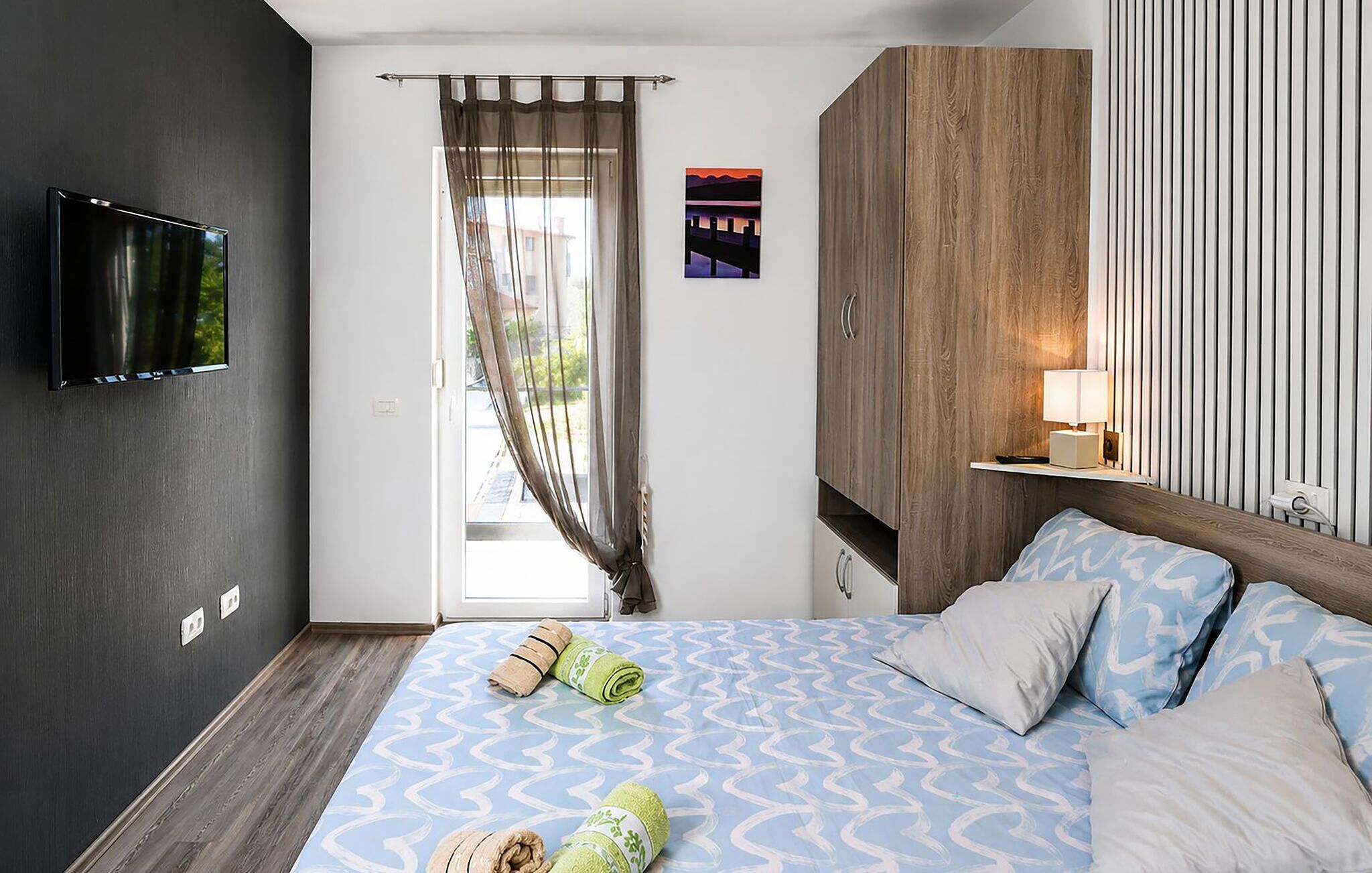 Une chambre à coucher avec un lit, une télévision, une fenêtre avec des rideaux, un tableau et une lampe de chevet.