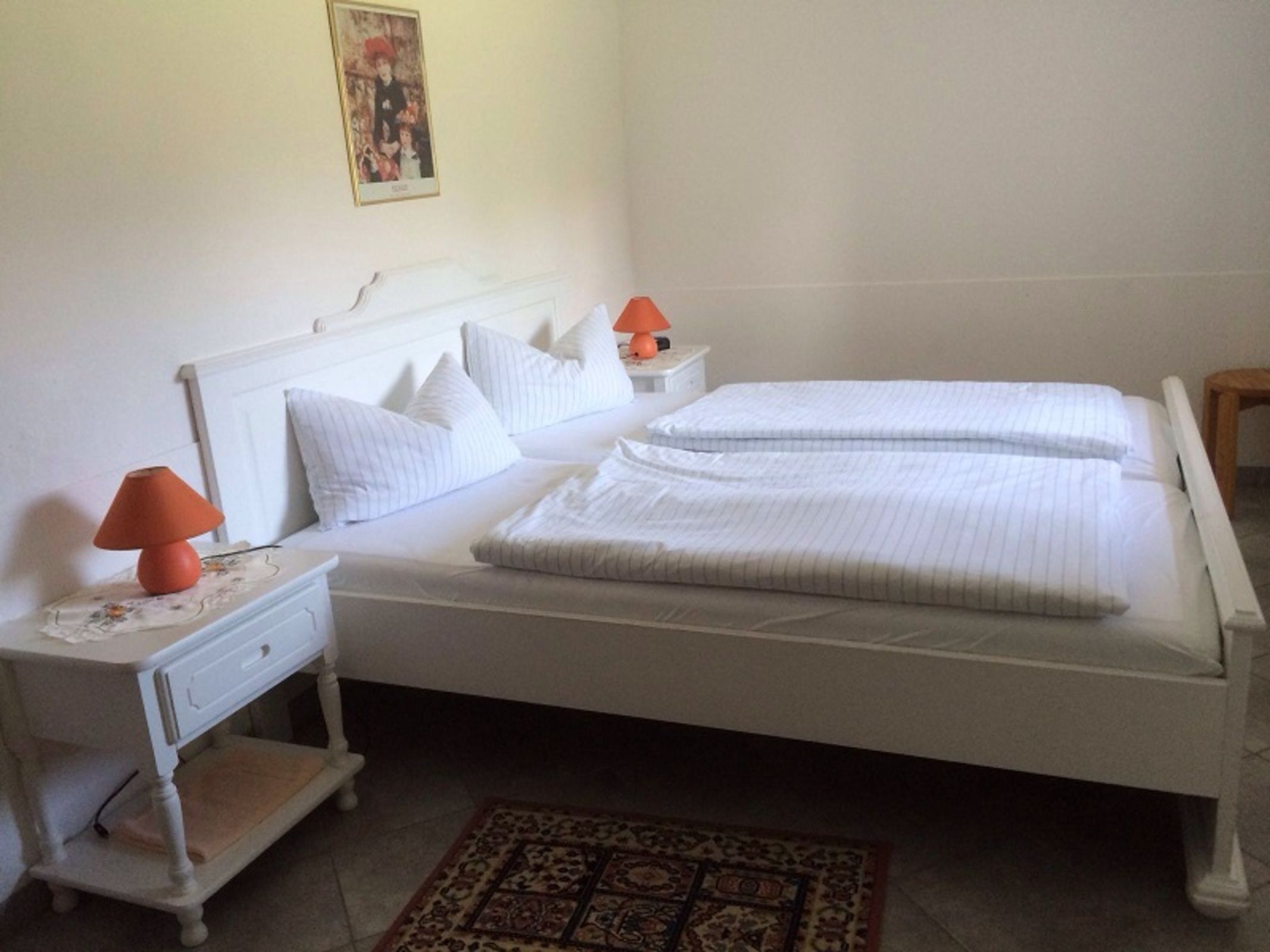 Un lit bien fait, avec du linge de lit blanc, une table de chevet avec une lampe et une petite commode de chevet également dotée d’une lampe.