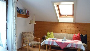 Una habitación acogedora con cabecera de madera, un sofá, una mesa de comedor con mantel, una silla, una ventana con vistas y una estantería.
