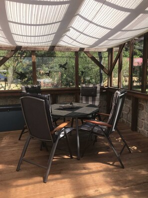 Un patio techado con una mesa redonda y sillas, ventanas amplias y vistas al verdor.
