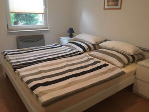 Ein Doppelbett mit gestreifter Bettwäsche, ein Nachttisch mit Lampe, eine Heizung und ein Fenster mit Jalousien.