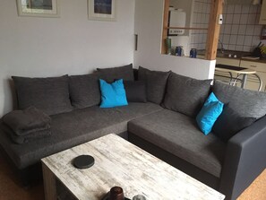 Ein graues Ecksofa mit blauen Kissen, ein weißer Couchtisch und eine Küchenzeile im Hintergrund.
