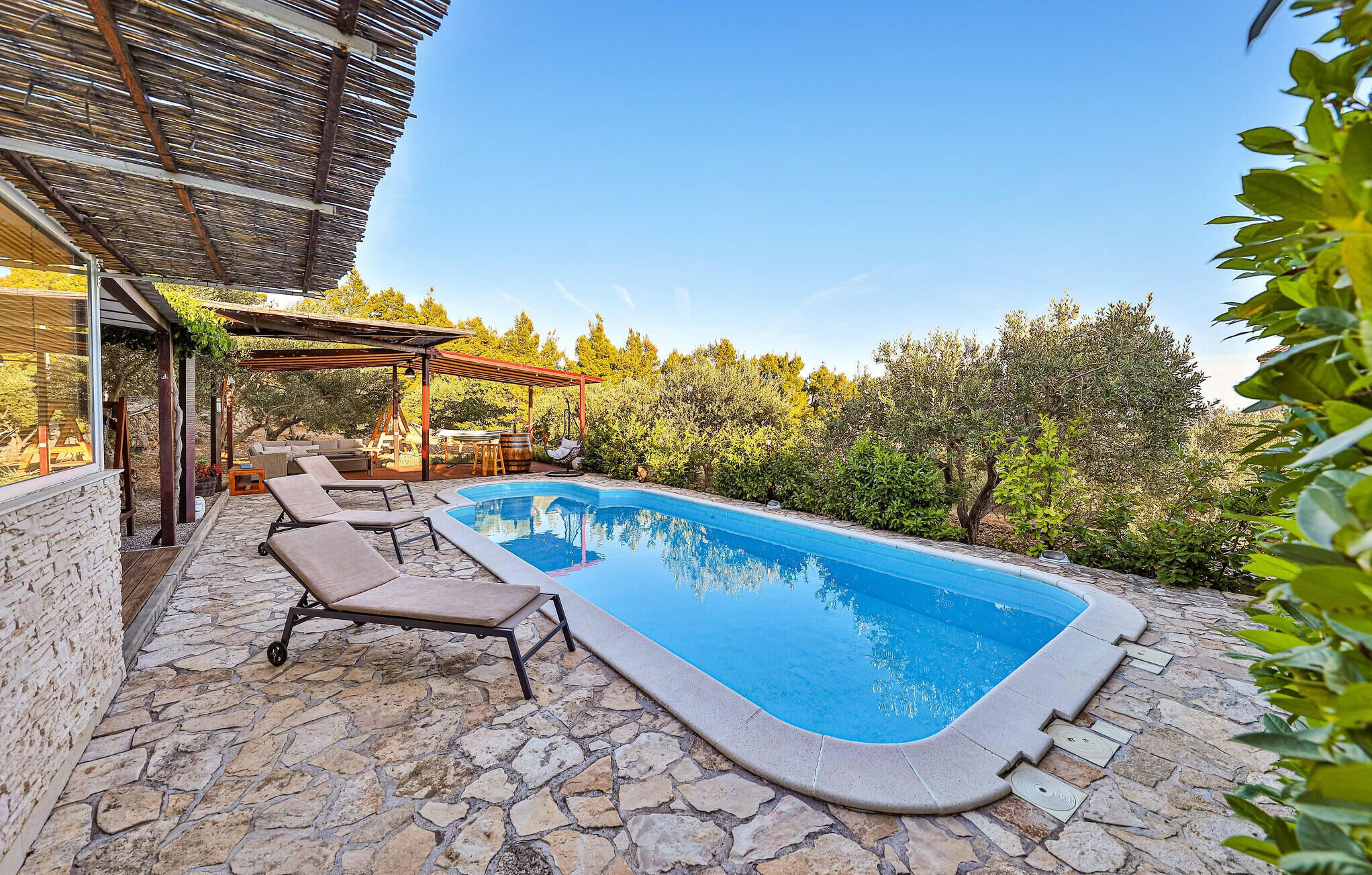 A swimming pool with lounge chairs, stone flooring, and a pergola with a view of trees.