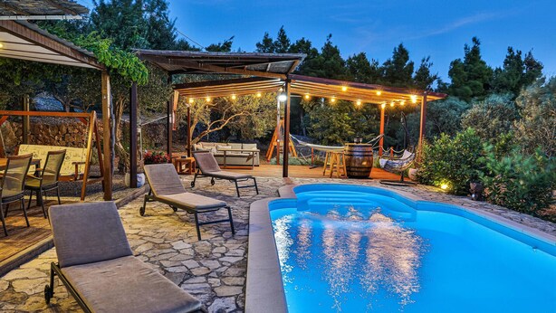 A backyard with a swimming pool, lounge chairs, a pergola with string lights, and a stone patio.