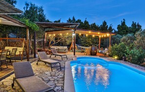 A backyard with a swimming pool, lounge chairs, a pergola with string lights, and a stone patio.