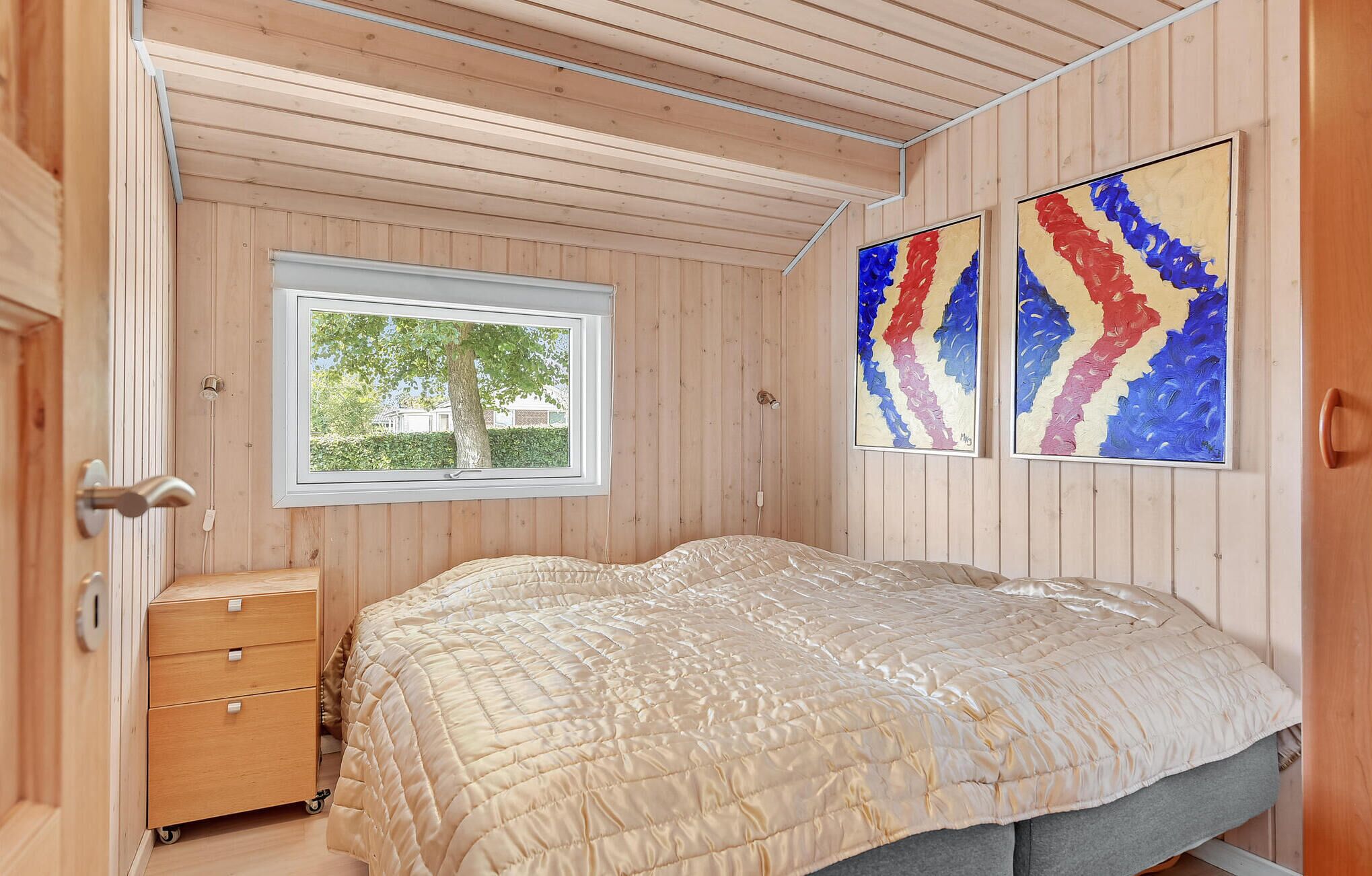 A bedroom with a wooden ceiling, a window with a view of greenery, a bed with a beige quilt, a wooden dresser, and two abstract paintings.