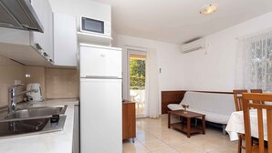 A compact kitchen with a refrigerator, microwave, and sink. There's a wooden table and a sofa in the living area, with a view of greenery outside. - Holiday apartment only 100 m from the sea (Baška)