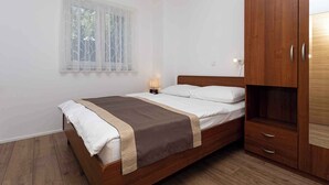 A bedroom with a wooden bed, a nightstand with a lamp, a wardrobe, and a window with sheer curtains. - Holiday apartment only 100 m from the sea (Baška)