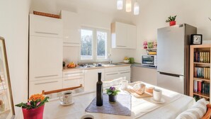 Een moderne keuken met witte kasten, een inbouwoven en een koelkast. Er is een raam dat natuurlijk licht biedt, en een eethoek met een tafel gedekt voor twee personen.