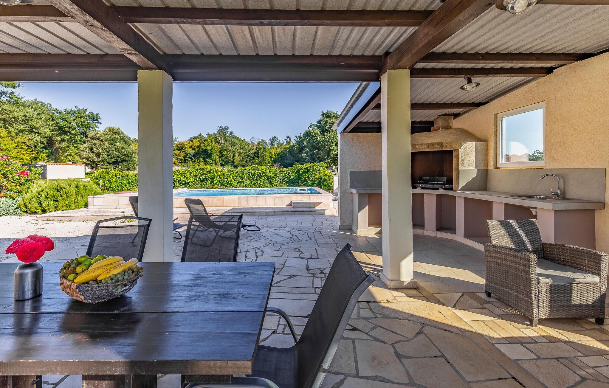 An outdoor patio with a table, chairs, a grill, and a pool in the background.