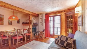 Un salon chaleureux avec un coin repas, des poutres en bois, une cheminée et une grande fenêtre ornée de rideaux rouges.