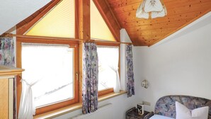 A bedroom with a sloped wooden ceiling, a large window with floral curtains, a bed with a floral headboard, and a small bedside table with a book. - gemuetliche Ferienwohnung (Kappl)