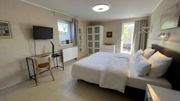 A hotel room with a bed, a desk, a chair, a TV, a window with curtains, and a door leading to an outdoor area.
