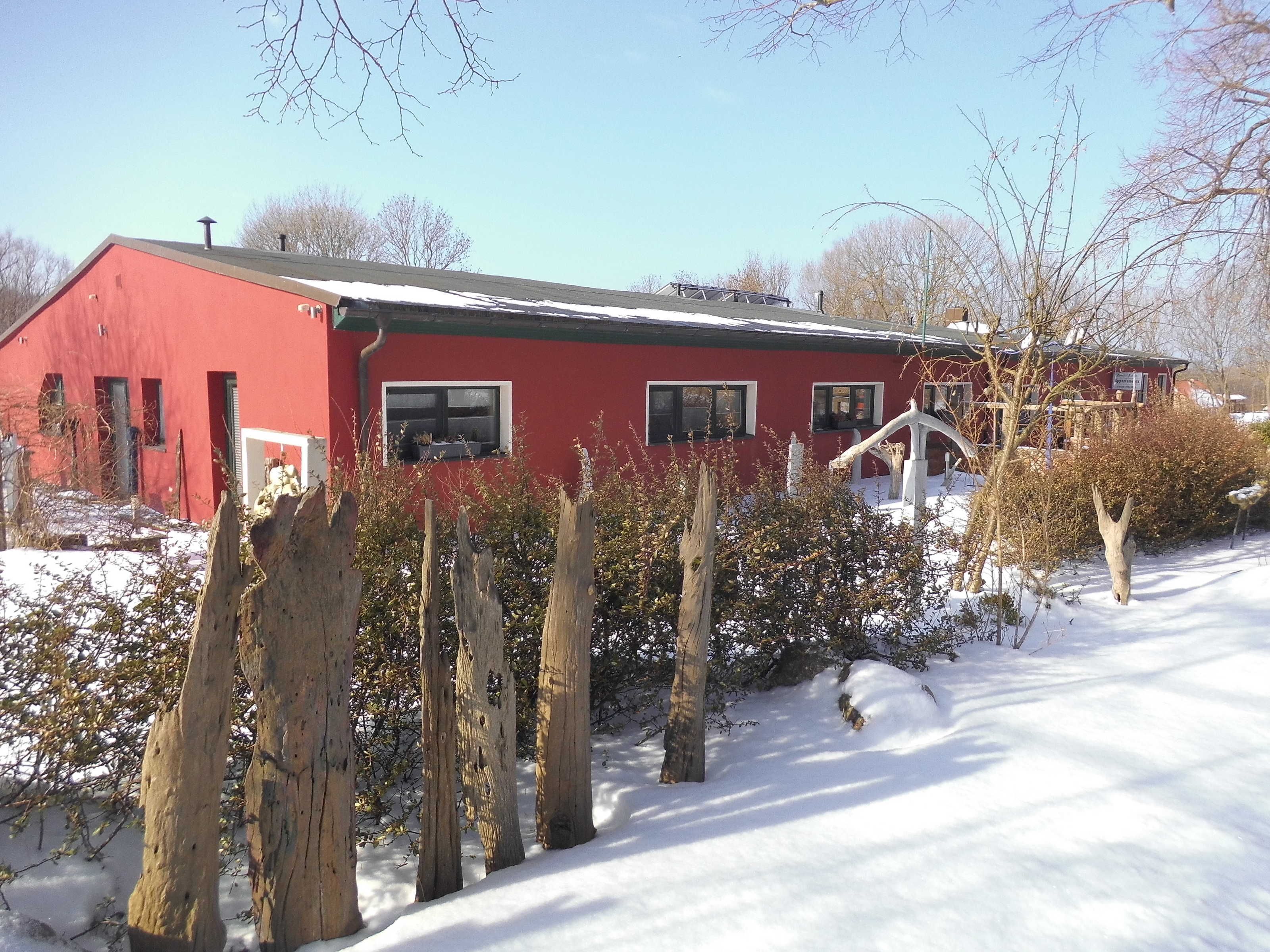 A red building with multiple windows, surrounded by snow-covered ground and bare trees.