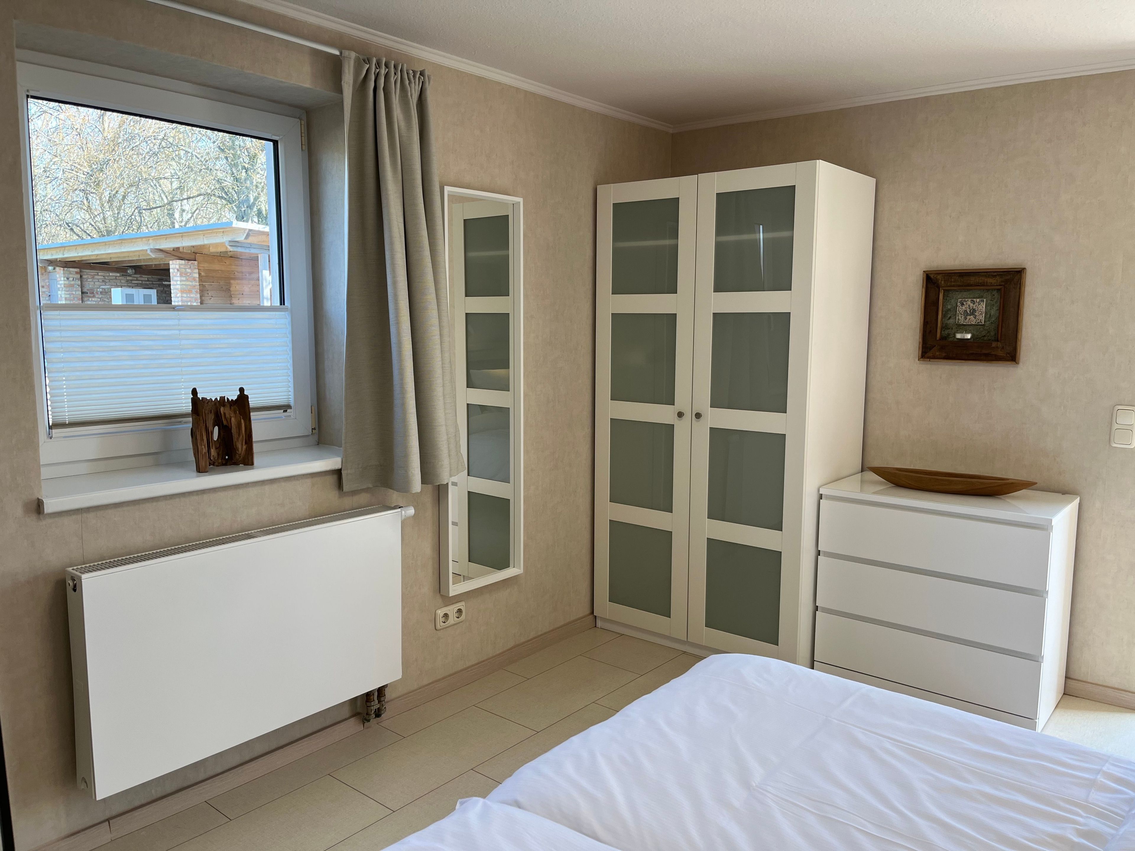 Ein Schlafzimmer mit einem Bett, einem Heizkörper, einem Fenster mit Vorhängen, einem Kleiderschrank und einem kleinen Tisch mit einer Schale.