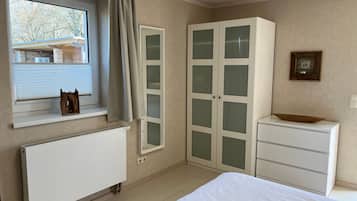 Ein Schlafzimmer mit einem Bett, einem Heizkörper, einem Fenster mit Vorhängen, einem Kleiderschrank und einem kleinen Tisch mit einer Schale.