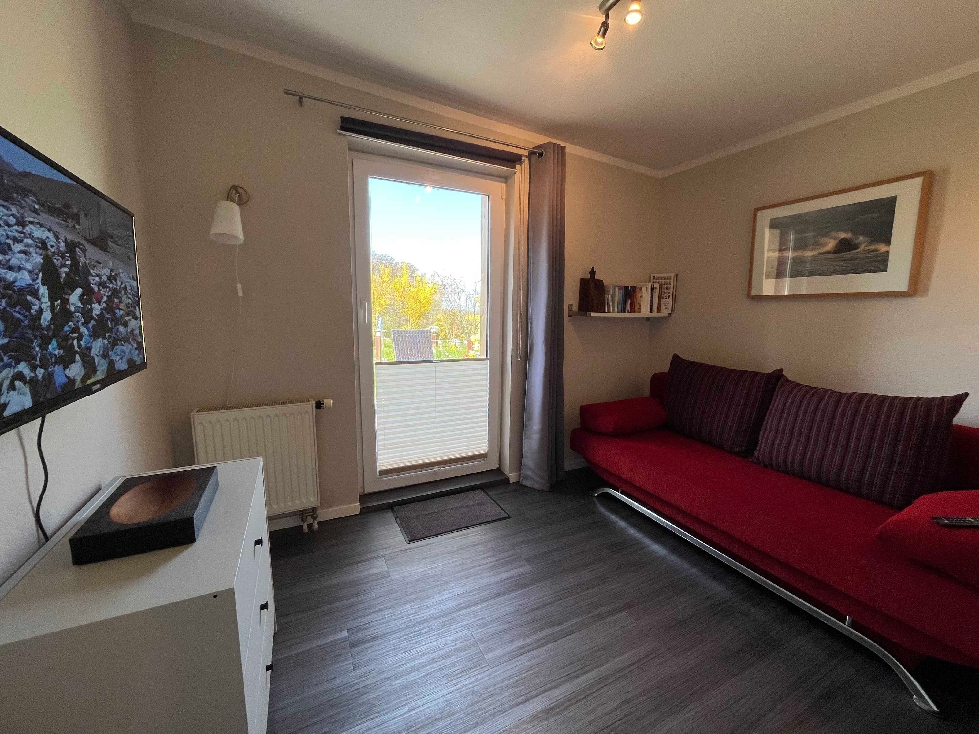 Et værelse med en rød sofa, et fjernsyn, en radiator, et vindue med gardiner og et billede i ramme på væggen.