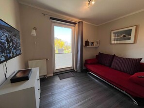 A room with a red sofa, a television, a radiator, a window with curtains, and a framed picture on the wall. - Findling - Kunsthof Salsitz (Lohme auf Rügen)