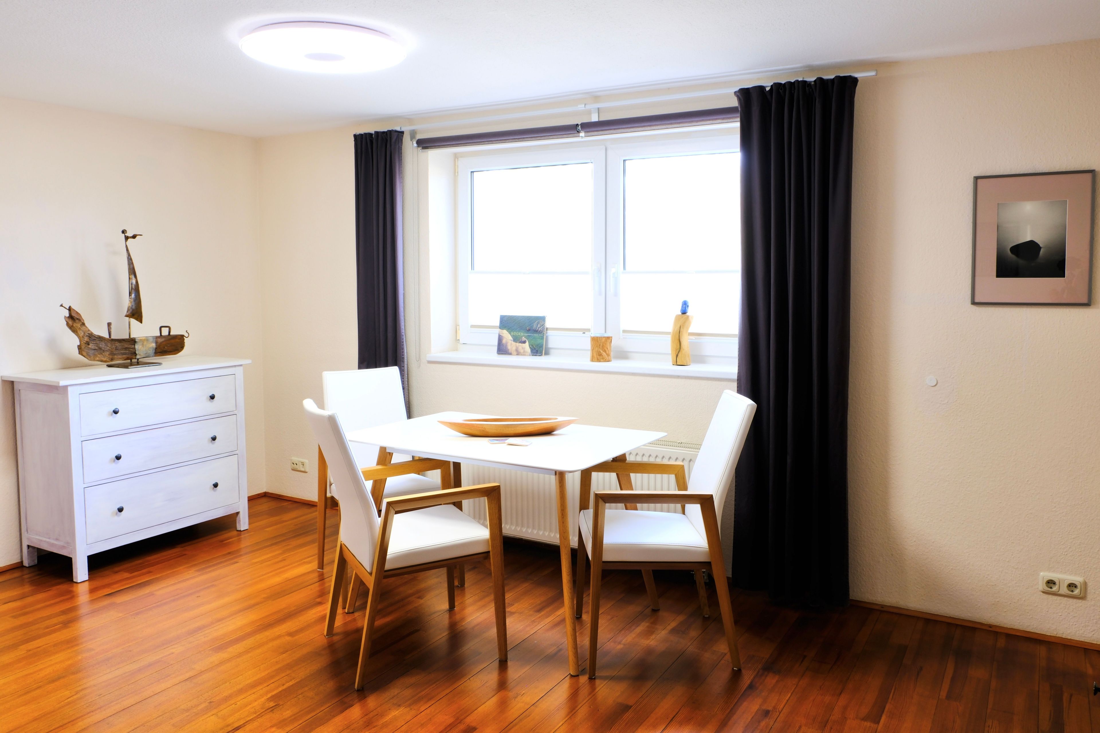 A dining area with a white table, wooden chairs, a white cabinet, and a window with black curtains.
