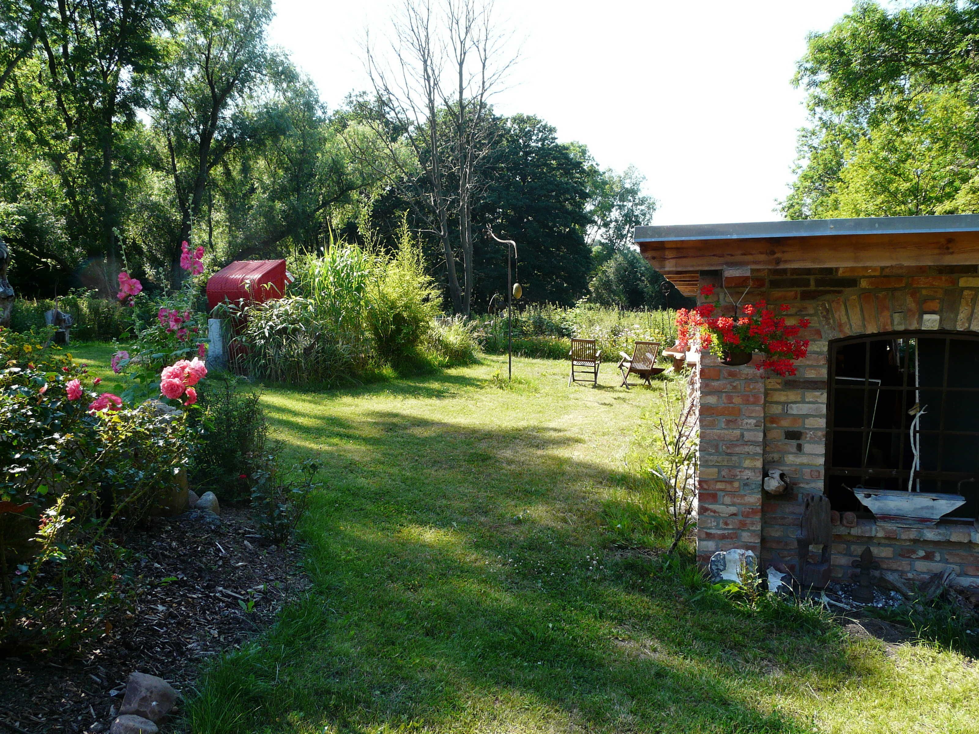 Ein Garten mit einer Ziegelstruktur, möglicherweise eine Terrasse oder ein Außenbereich mit Sitzgelegenheiten, umgeben von üppigem Grün und blühenden Pflanzen.