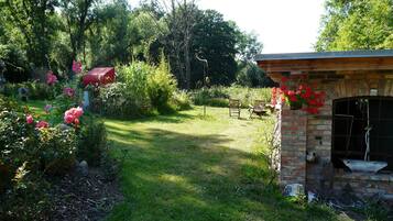 Ein Garten mit einer Ziegelstruktur, möglicherweise eine Terrasse oder ein Außenbereich mit Sitzgelegenheiten, umgeben von üppigem Grün und blühenden Pflanzen.