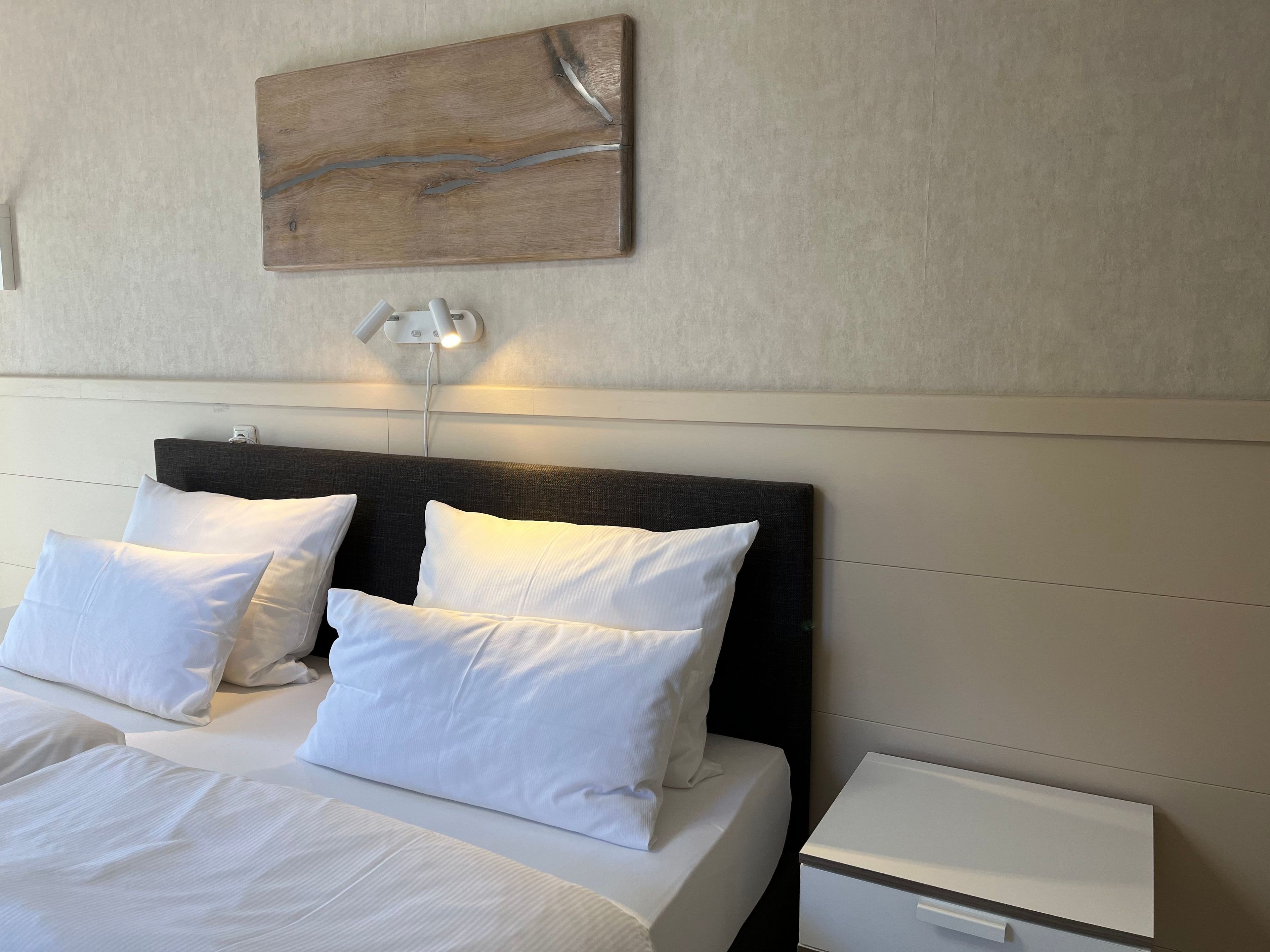Een hotelkamer met een bed, witte kussens, een nachtkastje, een wandlamp en een decoratief kunstwerk aan de muur.
