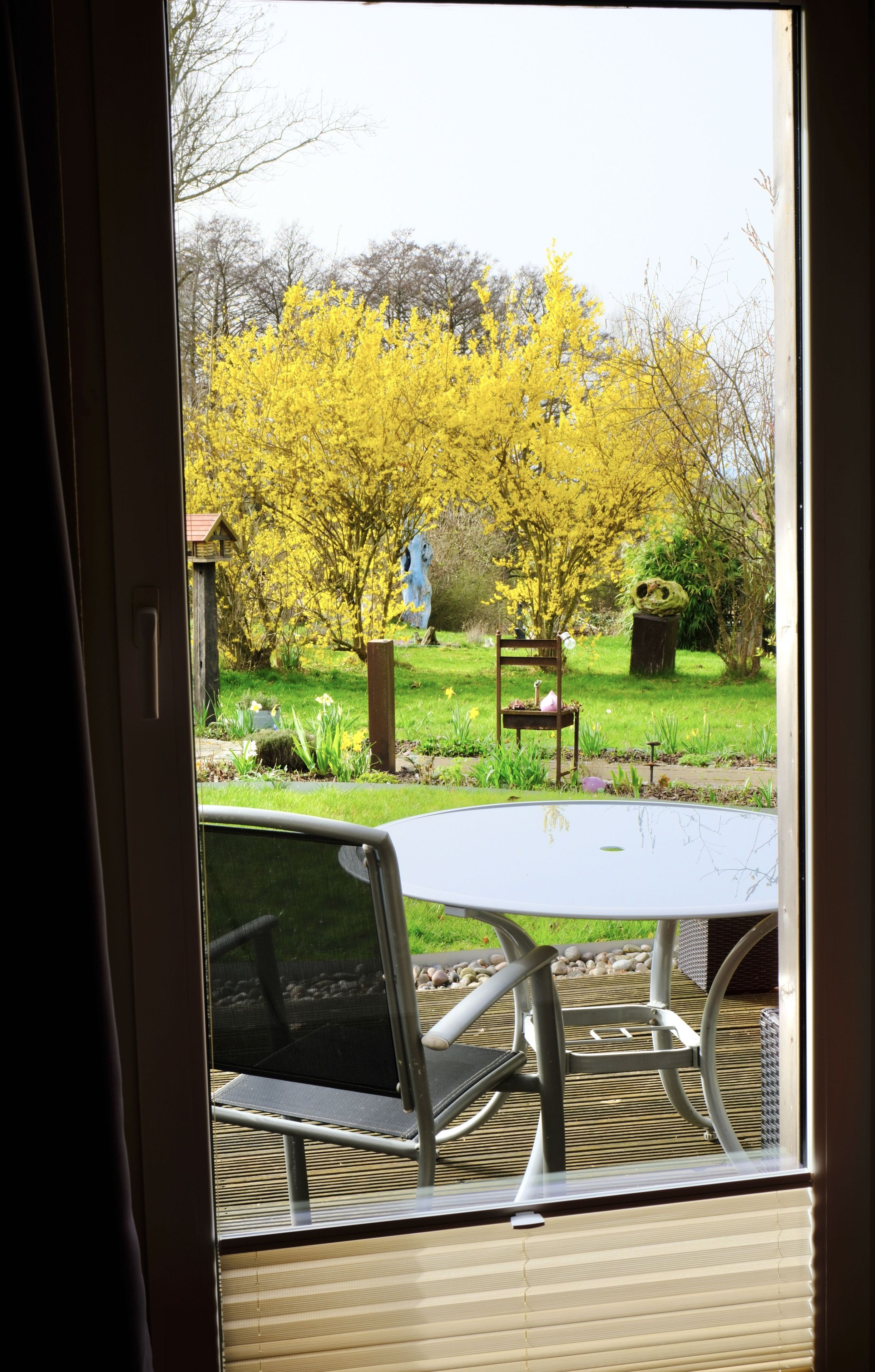 Blick durch ein Fenster, draußen Tisch und Stühle, ein Garten mit gelbem Laub und eine Statue im Hintergrund.