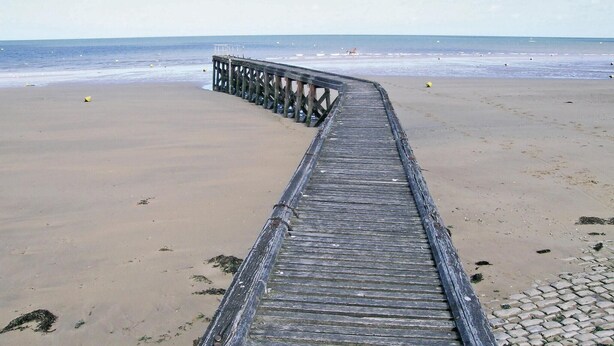 Une jetée en bois s’étendant sur une plage de sable, avec l’océan en arrière-plan.