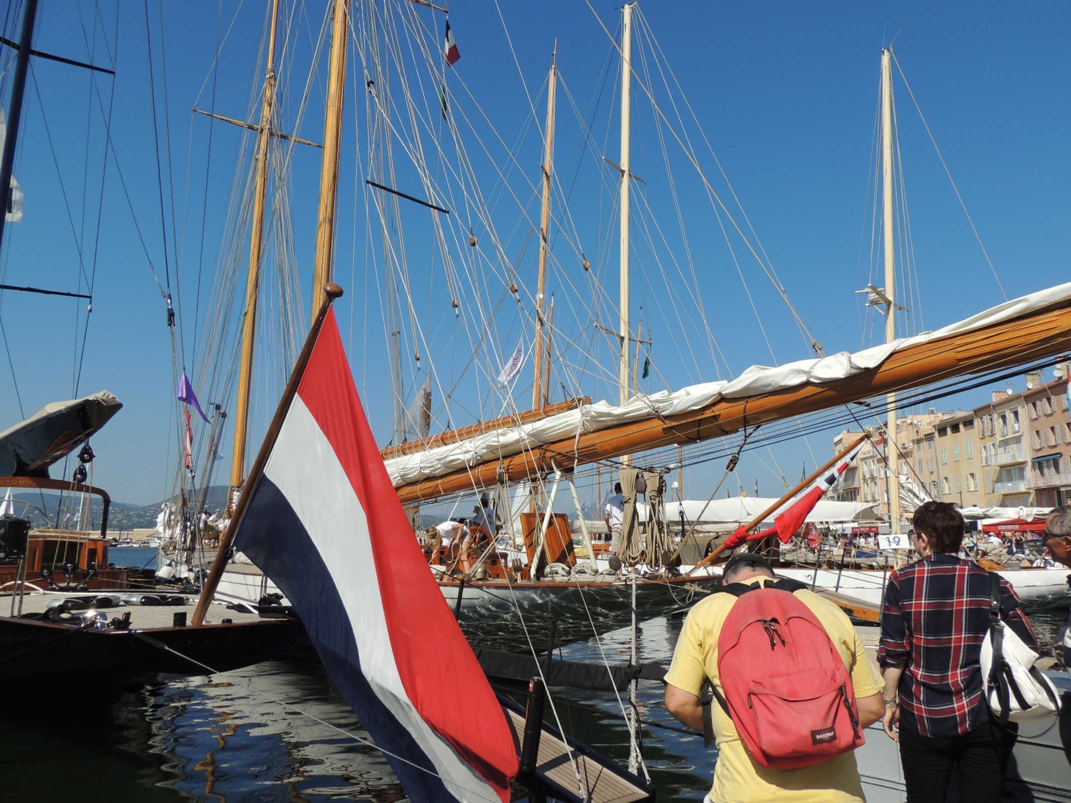 A marina with multiple sailboats docked, a flag, and people nearby.