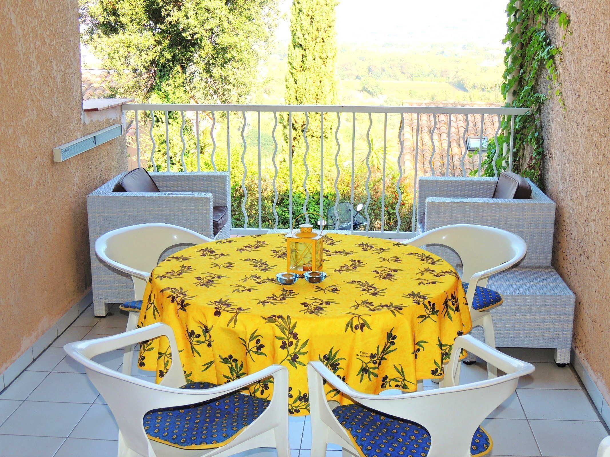 Un balcone con un tavolo rotondo ricoperto da una tovaglia gialla con motivo floreale, circondato da sedie bianche e cuscini blu.