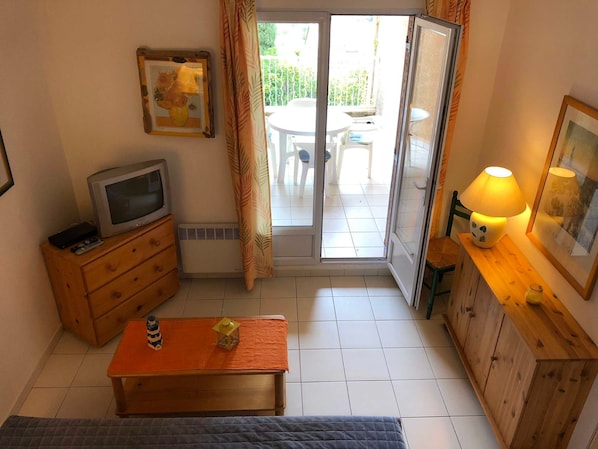A compact room with a television, a small table with a lamp, a wooden cabinet, and a sliding glass door leading to an outdoor area with a table and chairs.