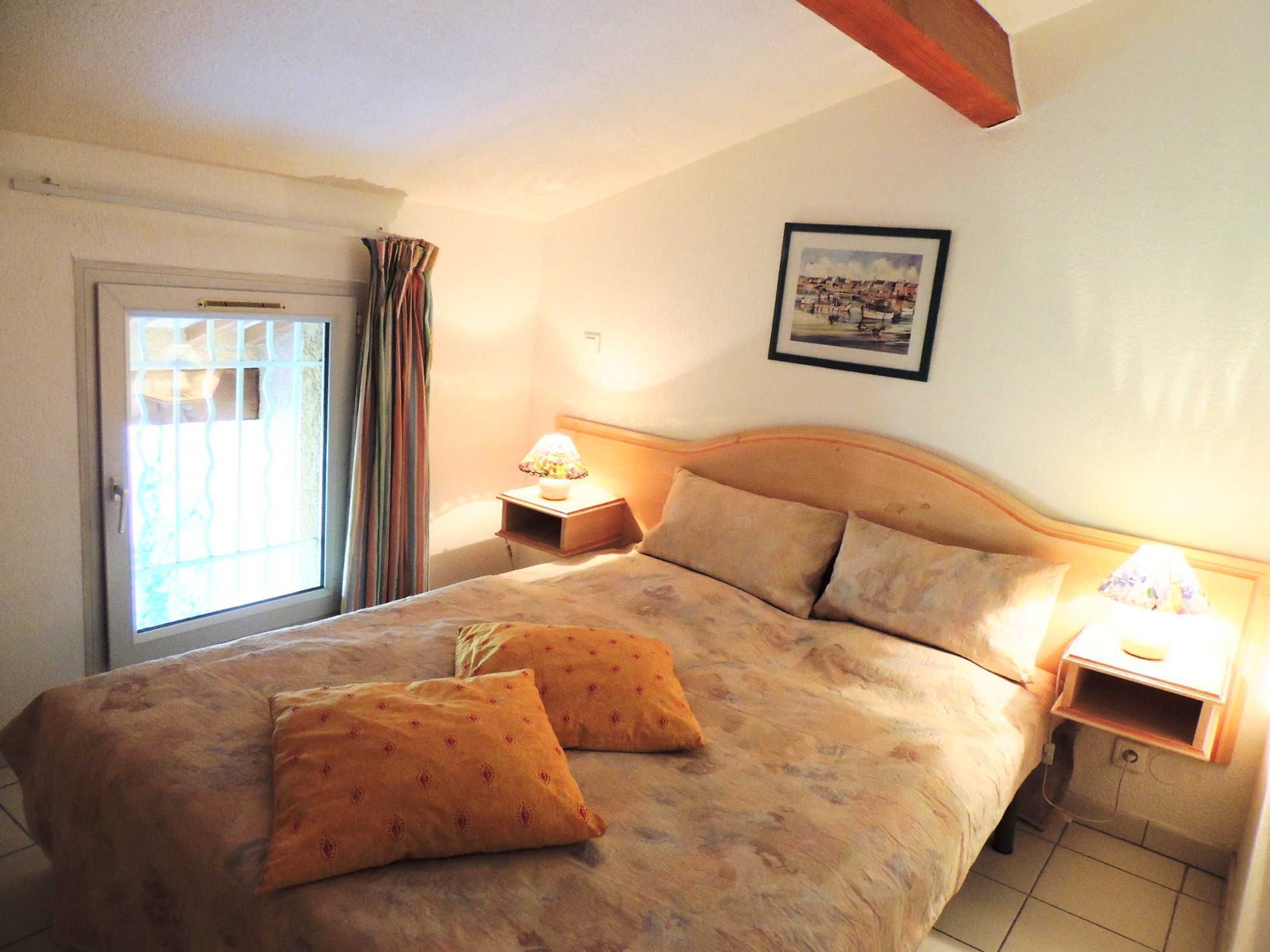 A bedroom with a bed, two pillows, a nightstand, a lamp, a window with curtains, and a framed picture on the wall.