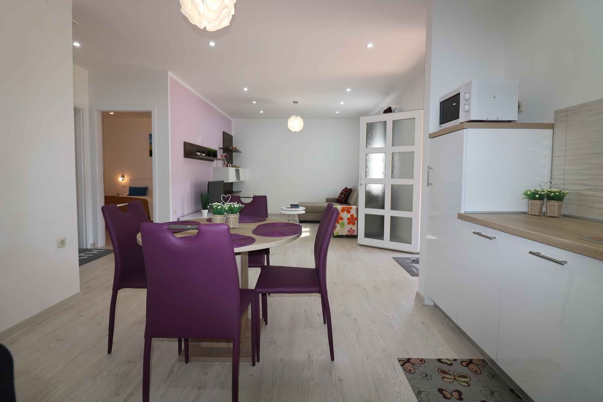 A compact living space with a dining area featuring purple chairs and a round table, a kitchenette with white cabinets, and a small seating area with a sofa and cushions.