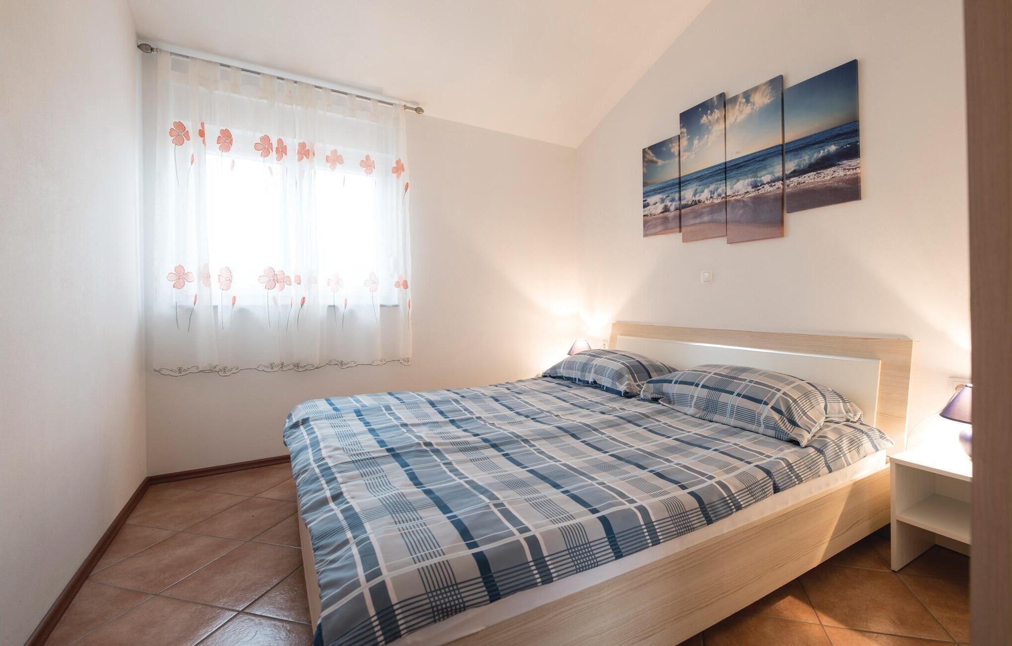 Une chambre à coucher avec un lit, une fenêtre ornée de rideaux à motifs floraux, une lampe de chevet et une œuvre d’art fixée au mur représentant une scène de plage.
