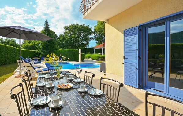 A poolside dining area with a table set for two, a pool, and a covered seating area with umbrellas.