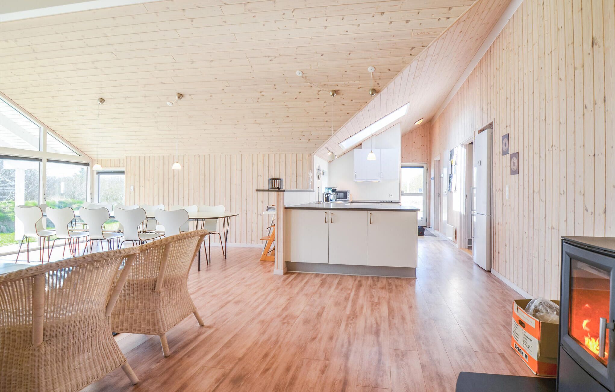 Een moderne keuken met houten vloeren, witte kasten en een centraal eiland. Er is een eethoek met witte stoelen en een rieten stoel. Een open haard is rechts zichtbaar.