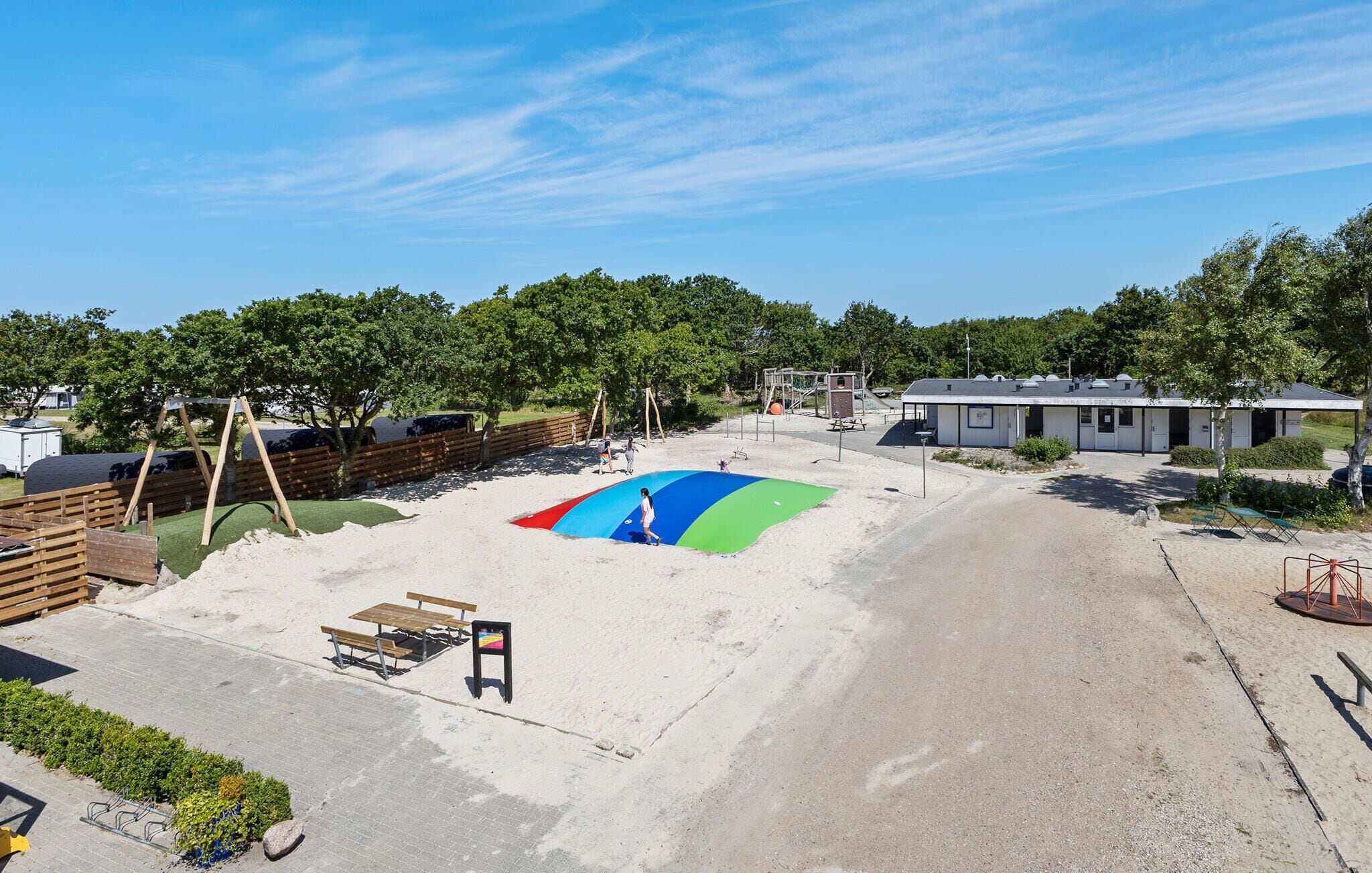 Ein Spielplatz mit Sandkasten und einem kleinen Gebäude, umgeben von Bäumen und einem Parkplatz.