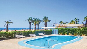 A swimming pool with a curved edge, surrounded by benches and palm trees, with a beach and ocean in the background. - Awesome apartment in Santa Susanna (Santa Susanna)