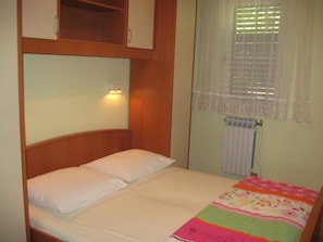 Una cama individual con colchón y almohadas blancos, cabecero de madera, lámpara de noche, radiador y ventana con cortina de encaje.